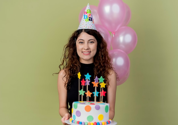 Young beautiful woman with curly hair in a holiday cap holding birthday cake smiling cheerfully happy and joyful standing over light wall