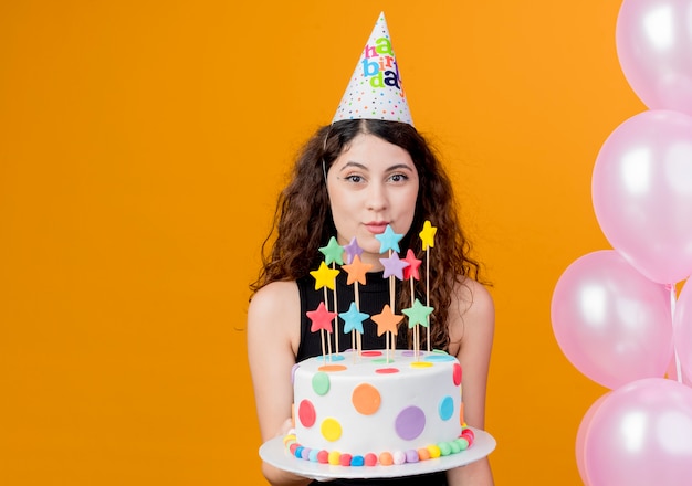 Free photo young beautiful woman with curly hair in a holiday cap holding birthday cake smiling cheerfully happy and joyful standing with balloons over orange wall