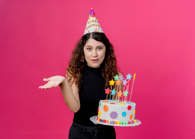 Young beautiful woman with curly hair in a holiday cap holding birthday cake  smiling with arm out as asking birthday party concept standing over pink wall