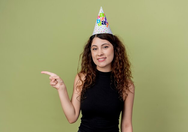 Young beautiful woman with curly hair in a holiday cap  pointign with finger to the side smiling cheerfully birthday party concept standing over light wall