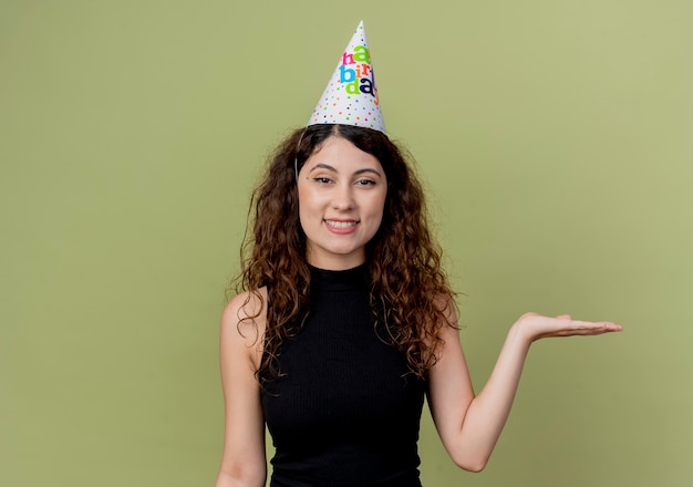 Young beautiful woman with curly hair in a holiday cap presenting with arm of hand smiling birthday party concept standing over orange wall