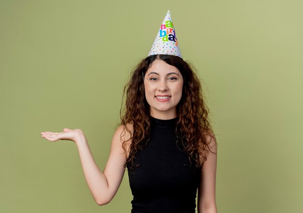 Young beautiful woman with curly hair in a holiday cap presenting with arm of hand smiling cheerfully birthday party concept standing over light wall