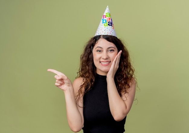 Young beautiful woman with curly hair in a holiday cap  smiling cheerfully pointing with finger to the side birthday party concept standing over light wall