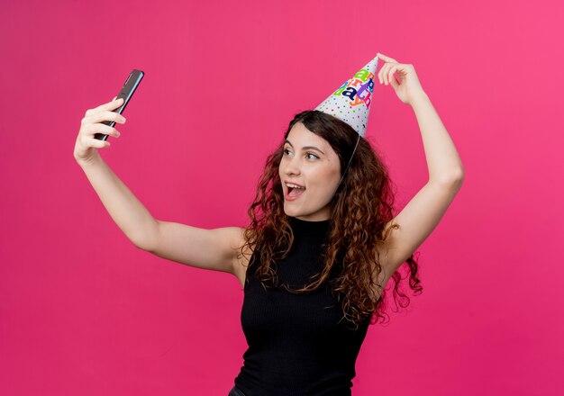 Young beautiful woman with curly hair in a holiday cap taking selfie smiling cheerfully birthday party concept  over pink