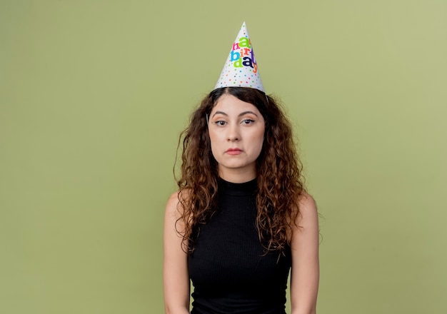 Young beautiful woman with curly hair in a holiday cap with sad expression on face birthday party concept standing over orange wall