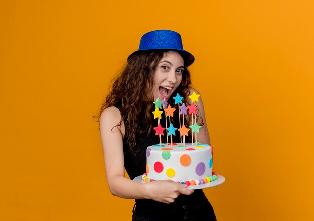 Young beautiful woman with curly hair in a holiday hat holding birthday cake happy and cheerful standing over orange wall