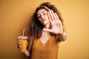 Free photo young beautiful woman with curly hair and piercing drinking healthy orange juice with open hand doing stop sign with serious and confident expression defense gesture