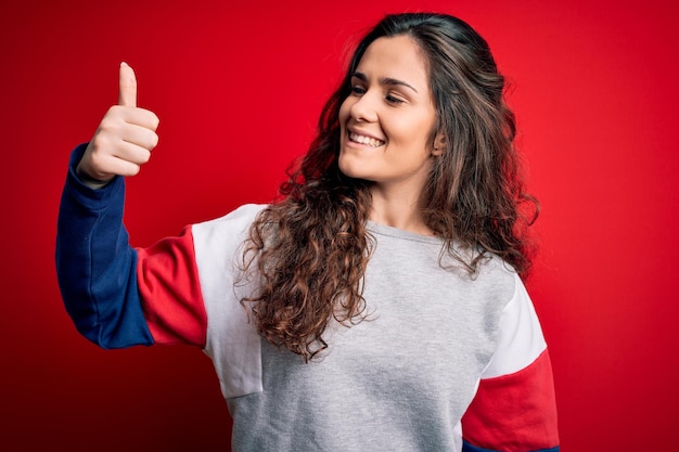 Young beautiful woman with curly hair wearing casual sweatshirt over isolated red background Looking proud smiling doing thumbs up gesture to the side