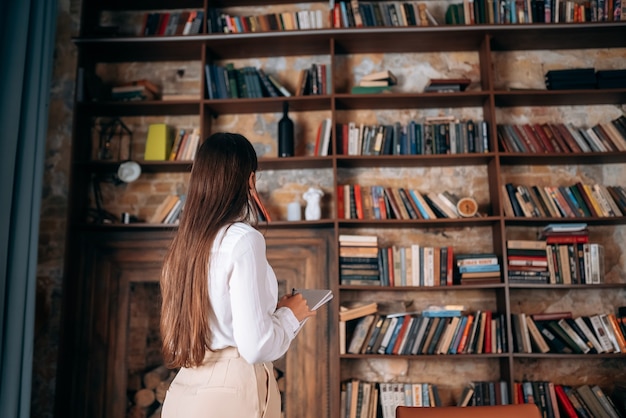 Young beautiful woman with glasses holding a notebook and looking at the bookshelf