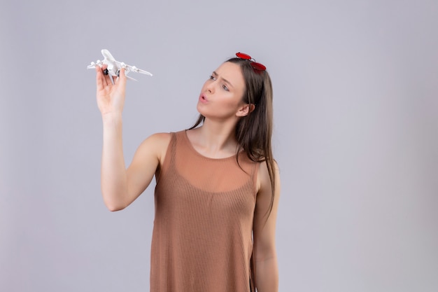 Free photo young beautiful woman with red sunglasses on head holding toy airplane looking playful and happy over white wall