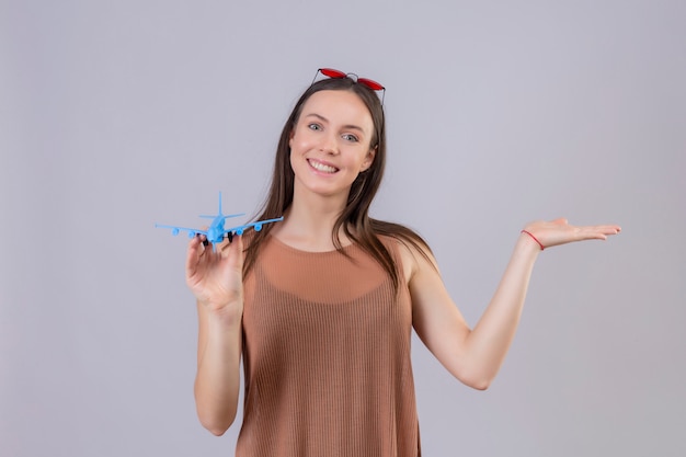 Young beautiful woman with red sunglasses on head holding toy airplane presenting with arm of hand smiling with happy face over white wall