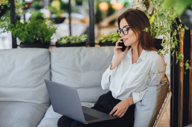 A young, beautiful woman works on a laptop on the summer terrace of her modern office and talks over the phone