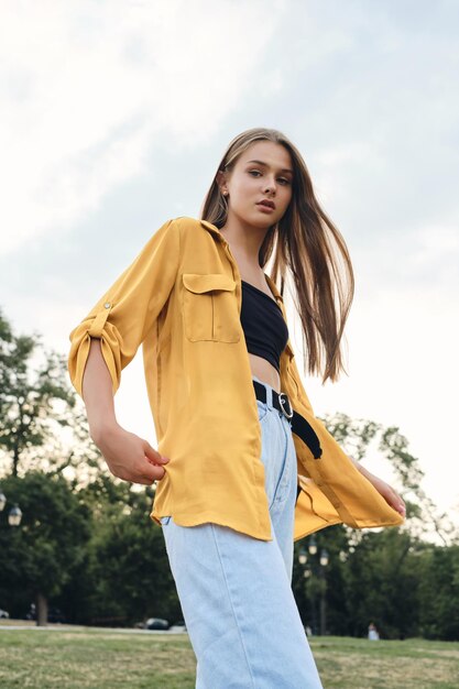 Young beautiful woman in yellow shirt and jeans dreamily looking in camera while standing on lawn in city park