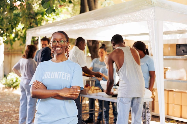 Young black woman with glasses stands outdoors, arms crossed, looking at the camera. Diverse group of volunteers supports a non-profit program dedicated to hunger relief and helping needy individuals.