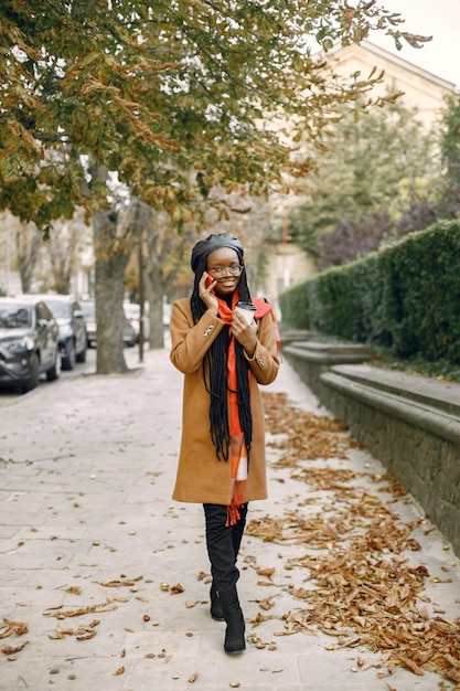 Free photo young black woman with a long locs hairstyles standing outside with a cup of take away coffee. woman wearing brown coat, orange scarf and black hat