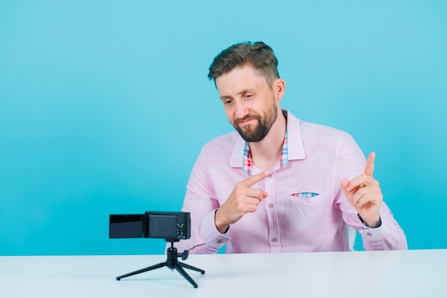 Free photo young blogger man is posing to his mini camera by pointing left with forefingers on blue background