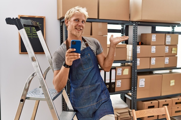 Young blond man using smartphone working at storehouse smiling cheerful presenting and pointing with palm of hand looking at the camera