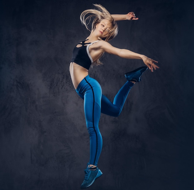 Young blonde ballerina in sportswear dances and jumps in a studio. Isolated on a dark background.