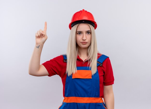 Young blonde engineer builder girl in uniform pointing up and looking  on isolated white wall