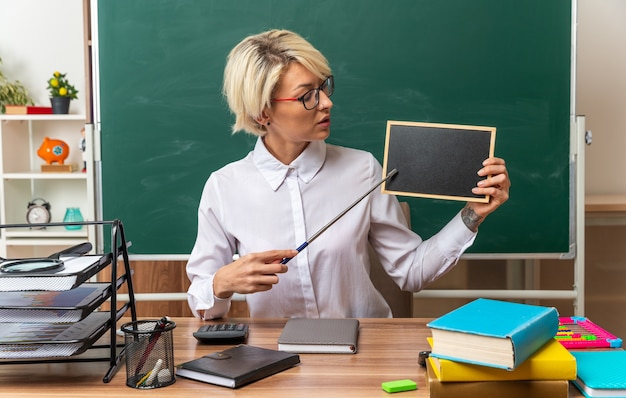 Young blonde female teacher wearing glasses sitting at desk with school tools in classroom showing mini blackboard looking at it pointing at it with pointer stick