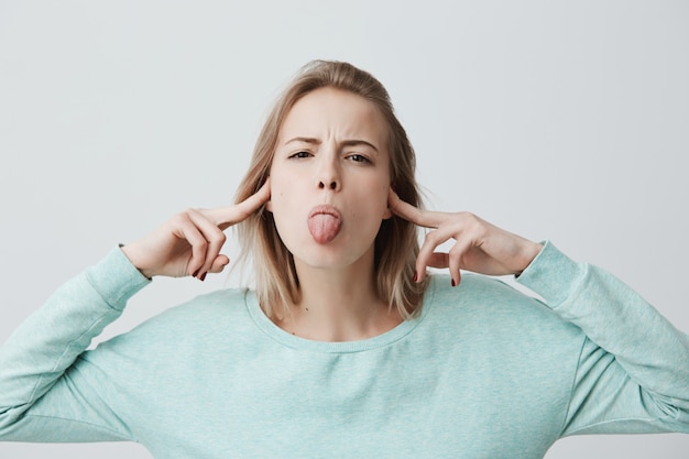 Young blonde female wears light blue clothes plugs ears with fingers, makes grimace, doesn t want to listen what she is told, sticks out her tongue in disregard. Face expression and body language