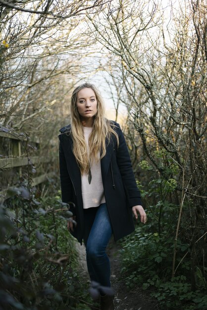 Young blonde female with a black coat standing on a pathway surrounded by leafless trees