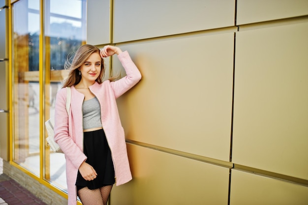 Young blonde girl in black skirt and pink coat posed against golden texture wall