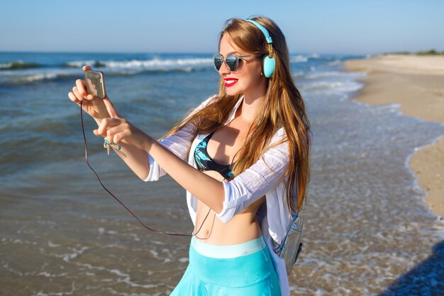 Young blonde girl having fun on the beach, bright hipster closing, vacation near ocean, listening music relaxing and making selfie on her phone.