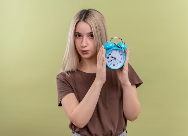 Young blonde girl holding alarm clock looking  on isolated green wall with copy space