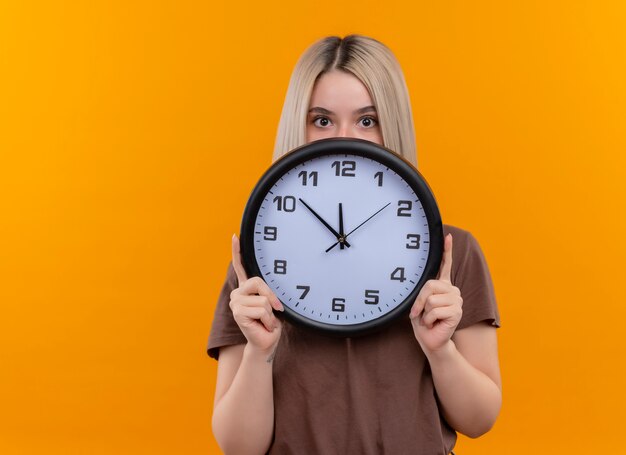 Young blonde girl holding clock hiding behind it on isolated orange wall with copy space