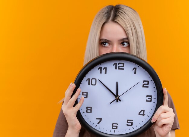 Young blonde girl holding clock hiding behind it looking at left side on isolated orange wall with copy space