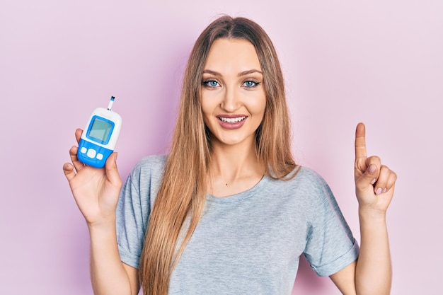 Young blonde girl holding glucometer device smiling with an idea or question pointing finger with happy face number one