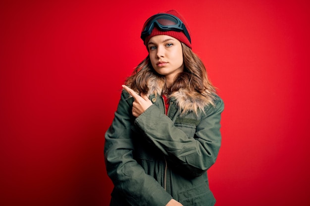 Free photo young blonde girl wearing ski glasses and winter coat for ski weather over red background pointing with hand finger to the side showing advertisement serious and calm face