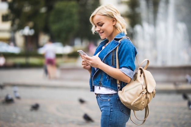 Young blonde girl woman phone in her hands on streetwalk square fontain dressed up in blue jeans suite with bag on her shoulder in sunny day