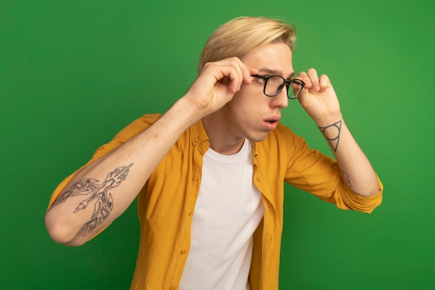 Free photo young blonde guy looking at side wearing yellow t-shirt and glasses isolated on green