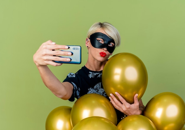 Young blonde party girl wearing masquerade mask standing behind balloons grabbing one of them doing kiss gesture taking selfie isolated on olive green background