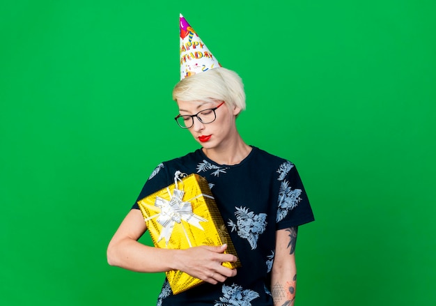 Free photo young blonde party woman wearing glasses and birthday cap holding and looking at gift box isolated on green wall with copy space