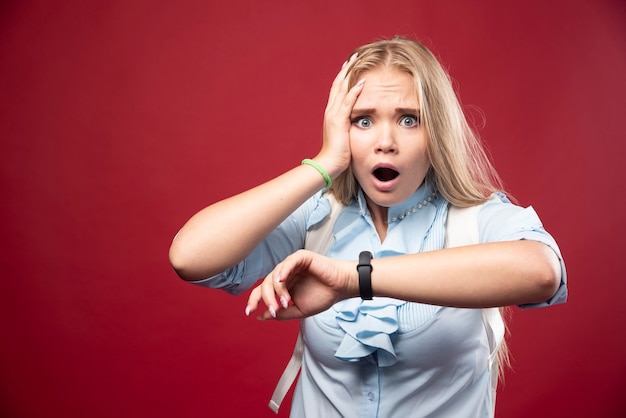 Young blonde student woman goes back to school, checks the time and gets stressed as she is late.