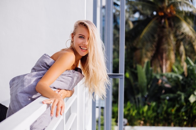 Free photo young blonde stylish european woman in blanket on tropical balcony meets sunrise in the morning.