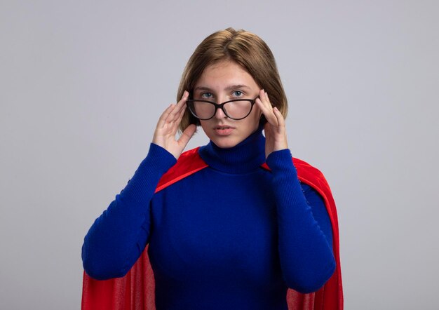 Young blonde superhero woman in red cape wearing glasses looking at front touching glasses isolated on white wall