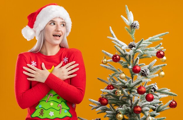 Young blonde woman in christmas  sweater and santa hat looking aside holding hands on her chest hapy and positive  standing next to a christmas tree over orange  background