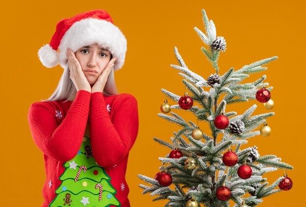 young blonde woman in christmas sweater and santa hat looking at camera with sad expression with arms on her cheeks standing next to a christmas tree over orange background