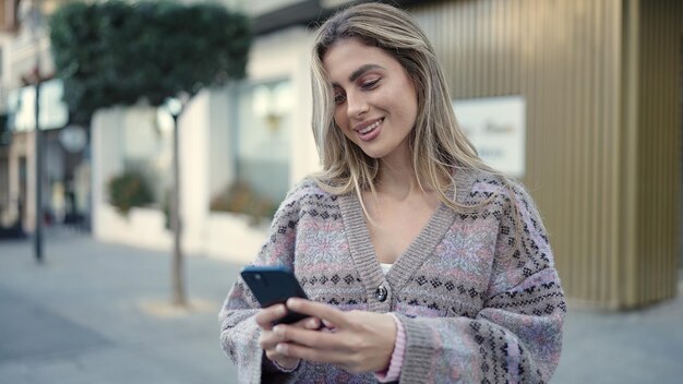 Young blonde woman smiling confident using smartphone at street