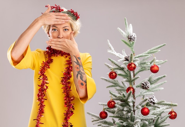 Free Photo young blonde woman wearing christmas head wreath and tinsel garland around neck standing near decorated christmas tree looking  keeping hands on forehead and on mouth isolated on white wall