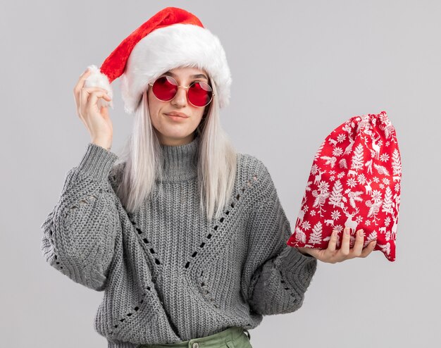 Young blonde woman in winter sweater and santa hat holding santa red bag with christmas gifts  happy and joyful looking aside standing over white wall