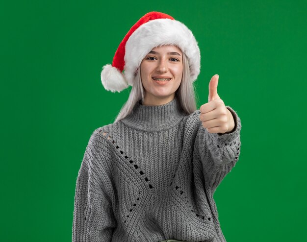 Young blonde woman in winter sweater and santa hat looking at camera smiling showing thumbs up standing over green background