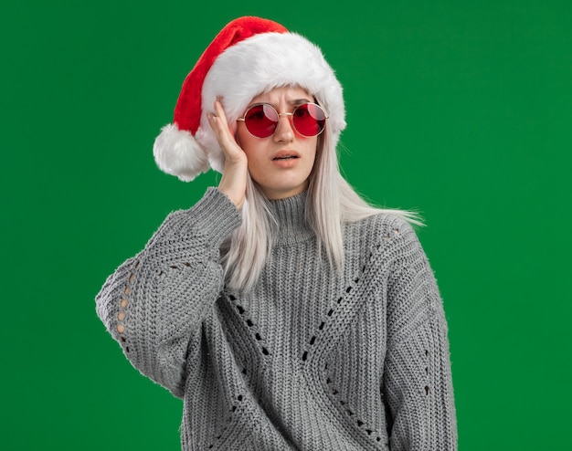 Young blonde woman in winter sweater and santa hat wearing red glasses looking confused with hand on her head  standing over green background