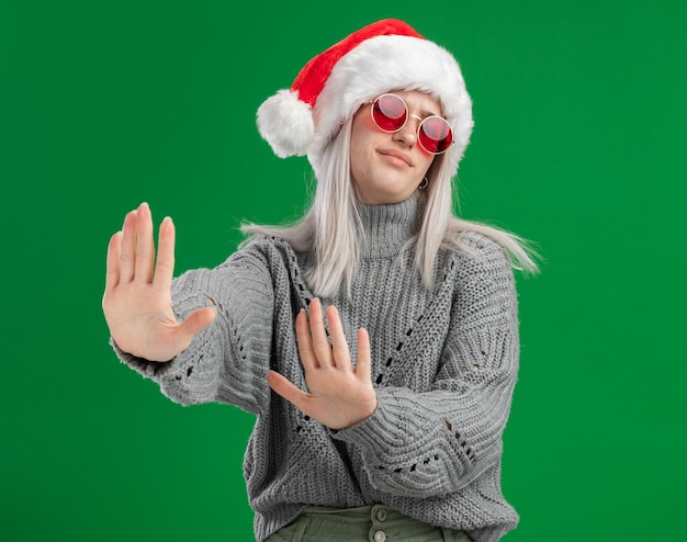 Free photo young blonde woman in winter sweater and santa hat wearing red glasses looking displeased making defense gesture with hands standing over green background