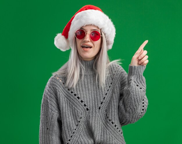 Young blonde woman in winter sweater and santa hat wearing red glasses looking surprised showing index finger having new idea  standing over green background