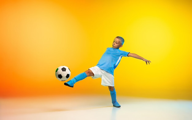 Young boy as a soccer or football player in sportwear practicing on gradient yellow studio background in neon light